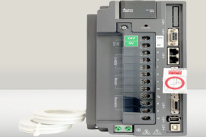 A PLC, showing a variety of ports and connectors, including power terminals, I/O terminals, and communication ports. The PLC is mounted on a metal backplate.