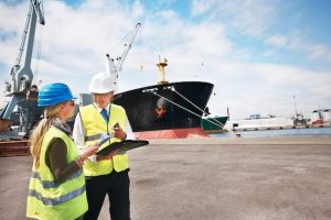 A man & a woman holding clipboard in front of cargo ship in ocean.