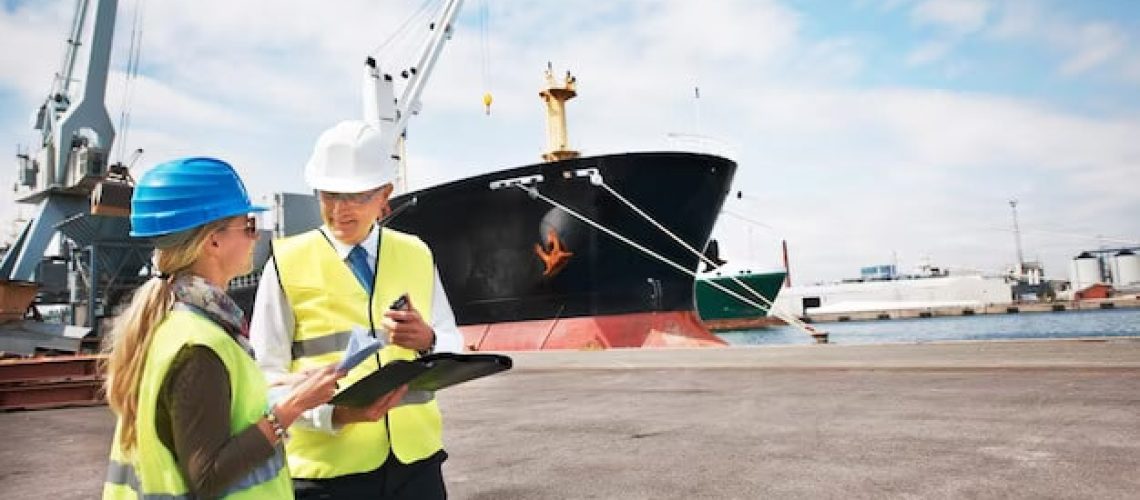 A man & a woman holding clipboard in front of cargo ship in ocean.